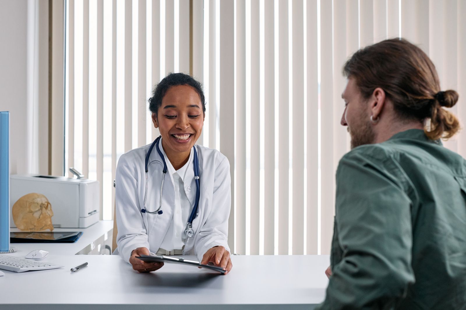 A doctor in a white coat smiling and talking with a patient seated across her desk