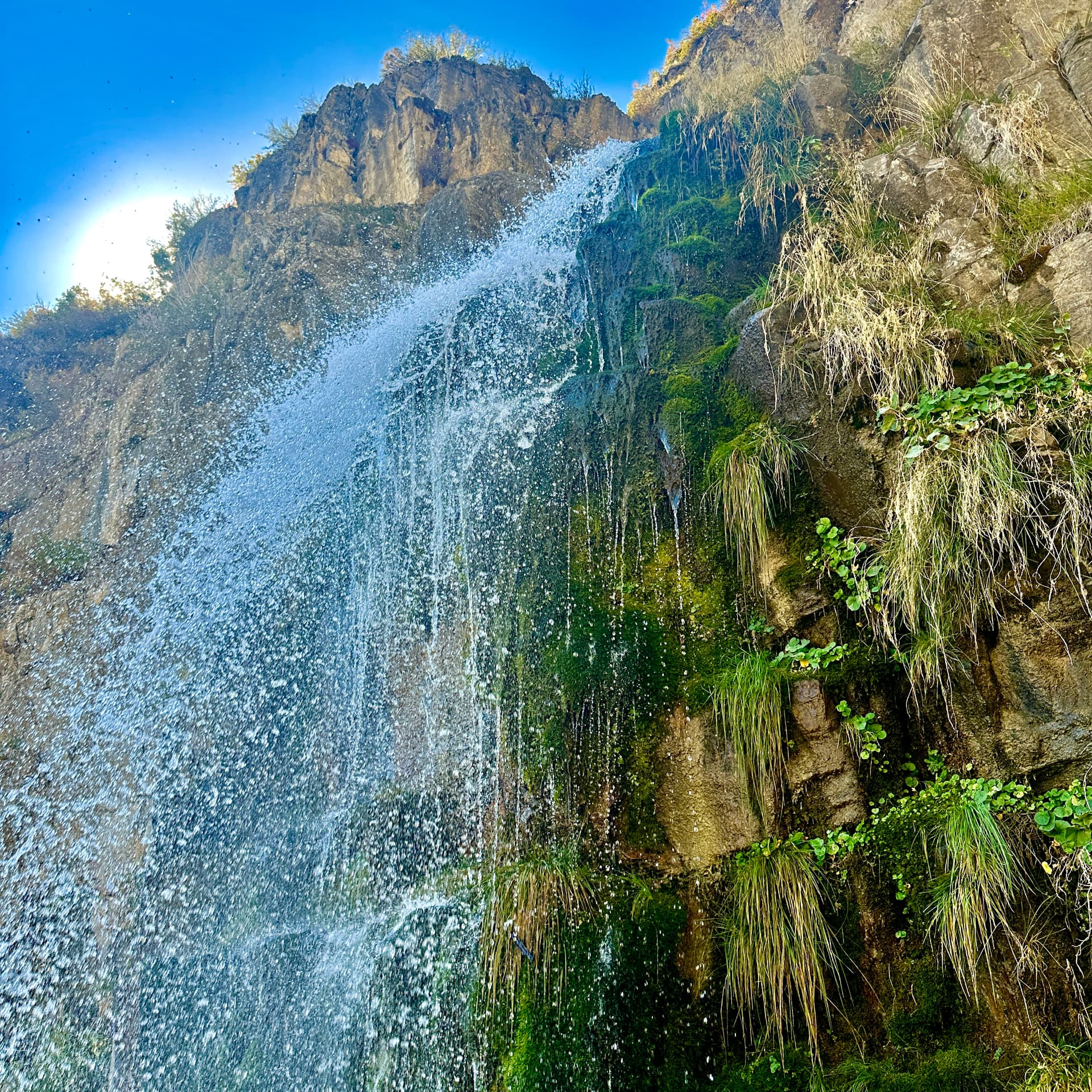A waterfall pouring over a mossy rock cliff on a Utah hiking trail under a blue sky