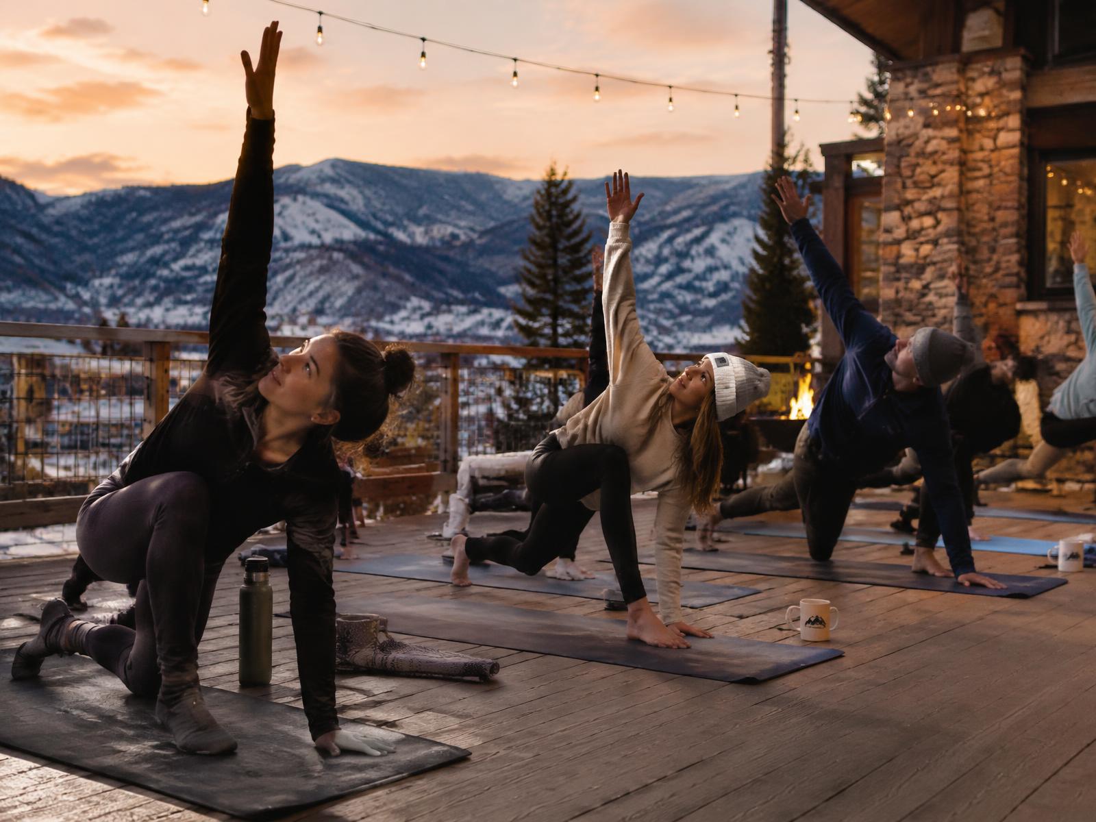 A group practicing yoga on a lodge deck with snowy mountains behind them